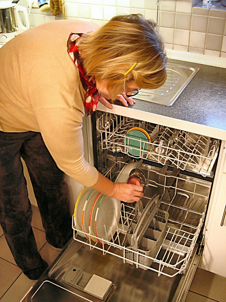 A woman leans over a dishwasher full of dishes.